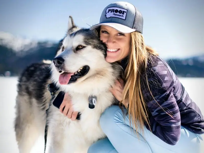 Mujer sonriente abrazando a un perro grande tipo Husky en un paisaje nevado, ambos irradiando felicidad y calidez.